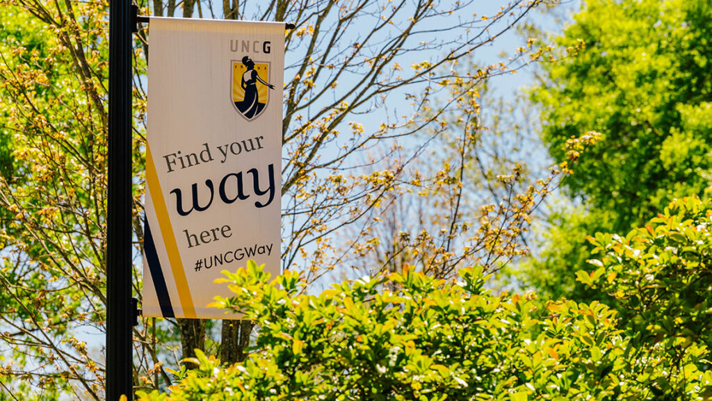 UNCG "Find Your Way Here" banner hangs from a pole in the trees.